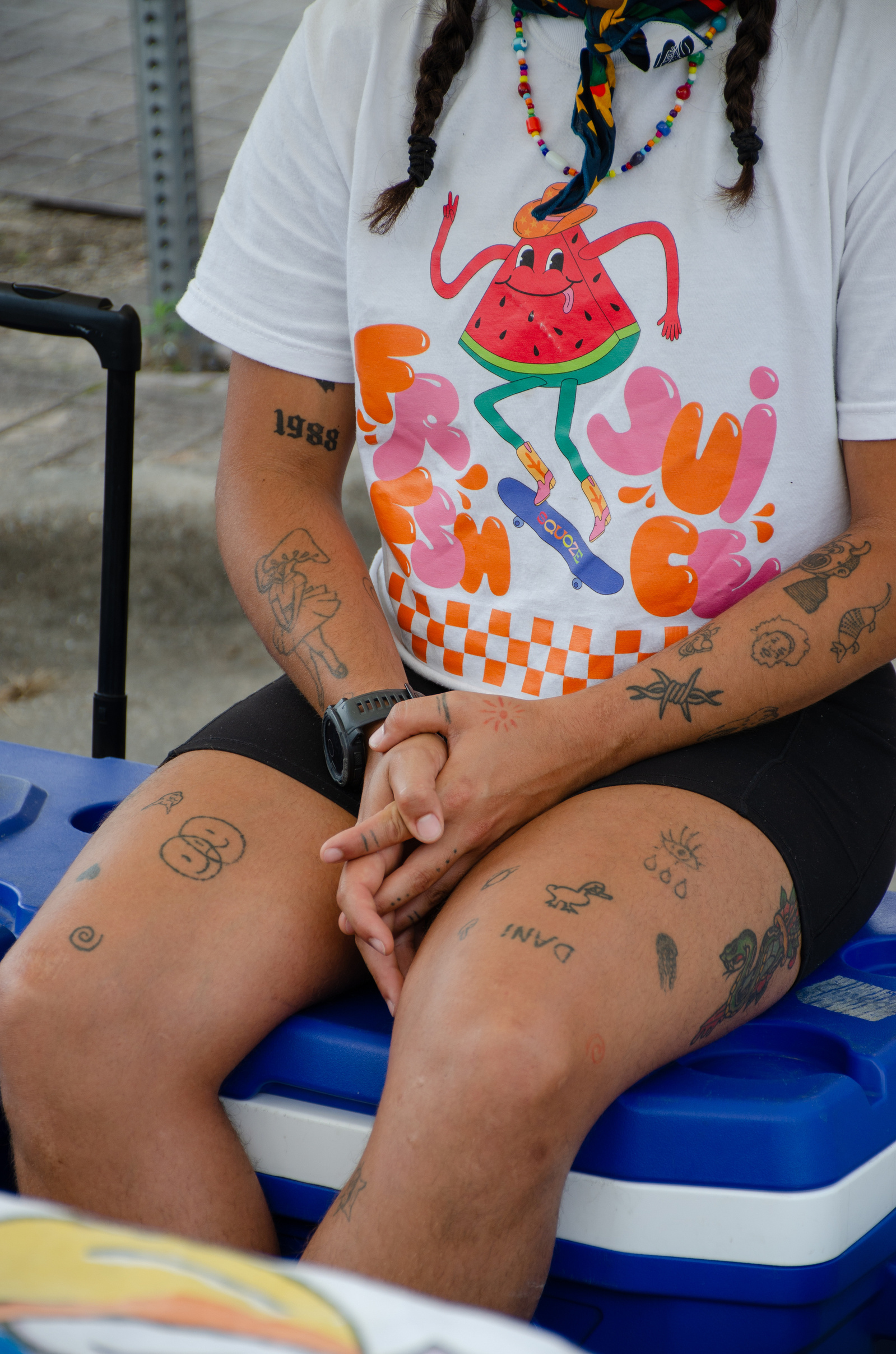 A portrait of summer skin mapped with ink, where each tattoo reads like a page from a personal zine. The watermelon tee and woven beads nod to humor and youth, but the stillness of the hands suggests something quieter—an intermission between stunts and st
