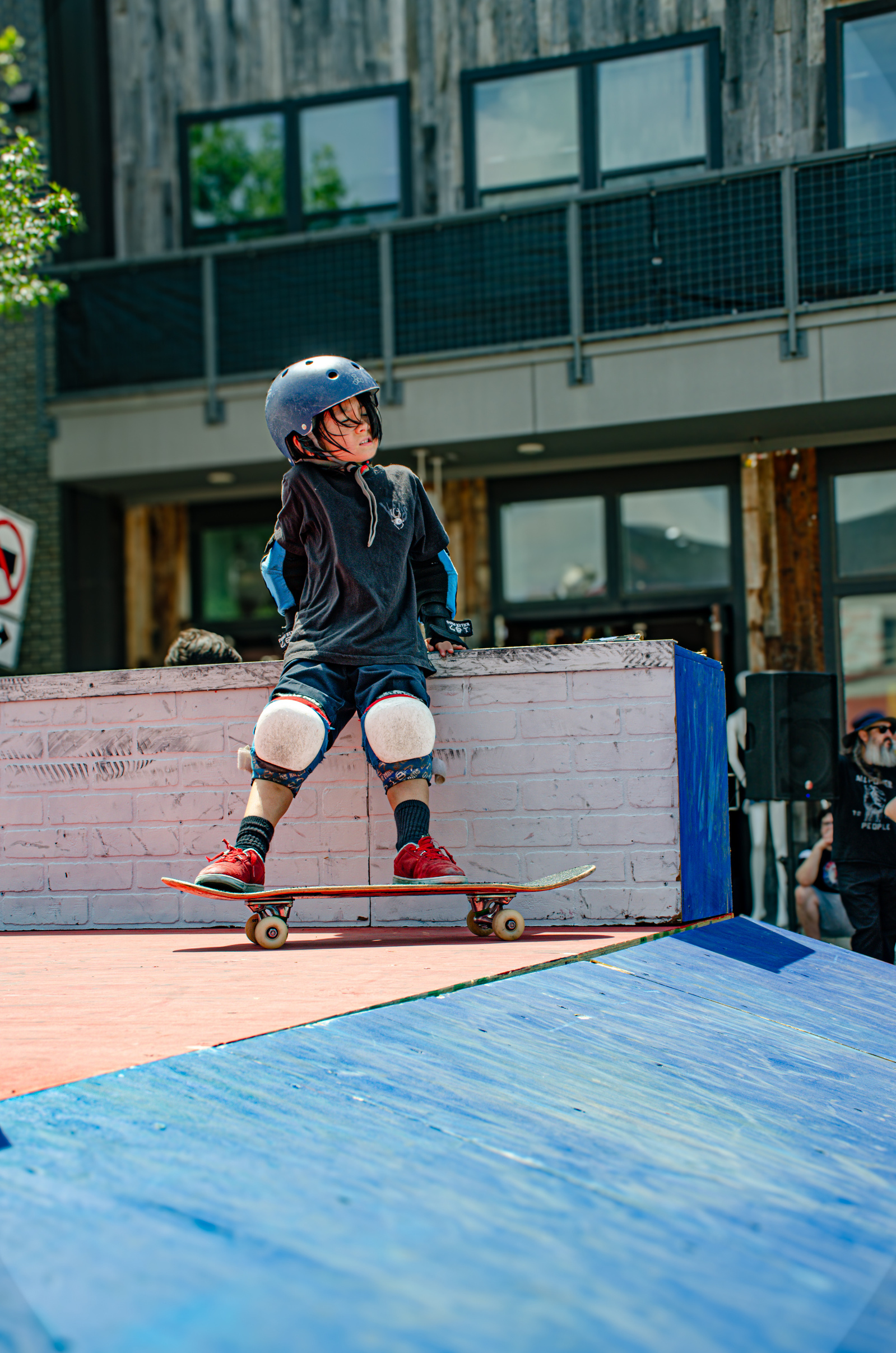 Perched and padded, a young skater rides the edge of courage. There’s no rush to drop in—just curiosity, heat, and a brick ledge that’s both obstacle and invitation.