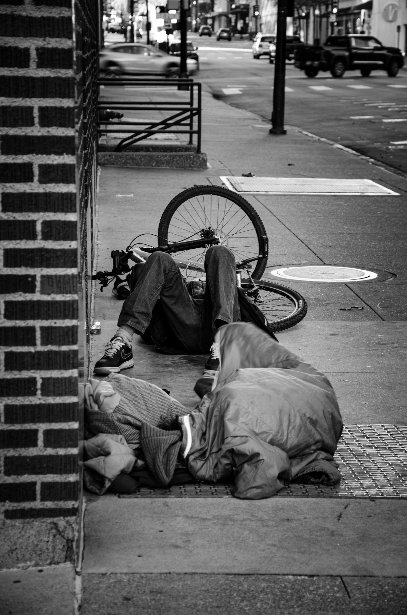 A body sprawled in stillness, a wheel forever turning. Here, the street offers rest, not respite. The city spins on, indifferent. He remains—part of the pavement, part of the picture.