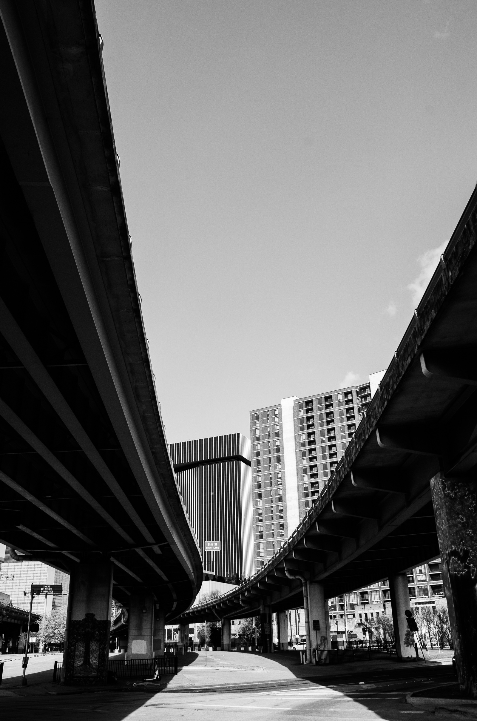A corridor of concrete arcs upward, framing a sliver of sky between rising buildings—an accidental cathedral shaped by infrastructure. This image considers how cities impose form without asking permission.