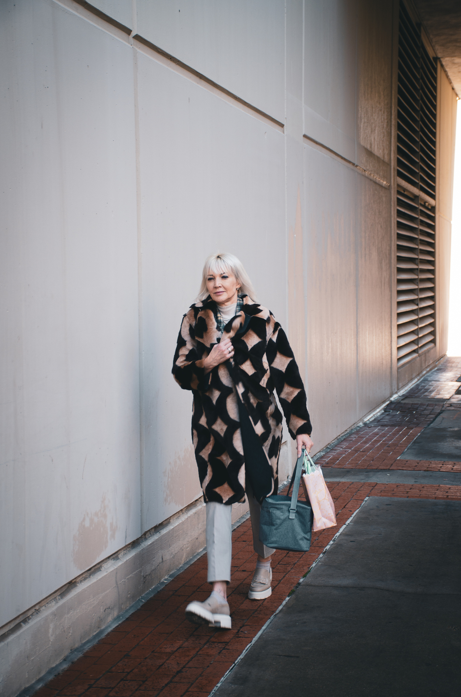 A woman strides with confidence, her coat a pattern of bold defiance. Between shadows and blank walls, she claims her space. Grace and gravity coexist here—style as armor, and movement as statement.
