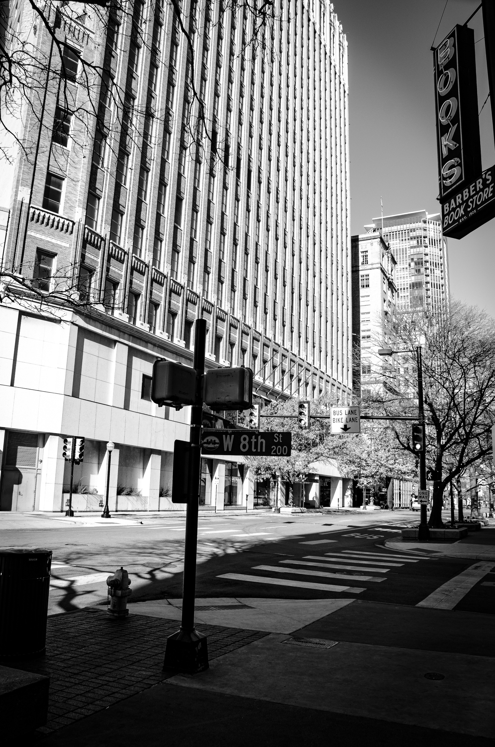 Morning light casts long shadows across the historic Book District, where Art Deco architecture tells stories of cultural heritage. The stark black and white composition emphasizes the geometric interplay of light and shadow across the canyon of W 8th.