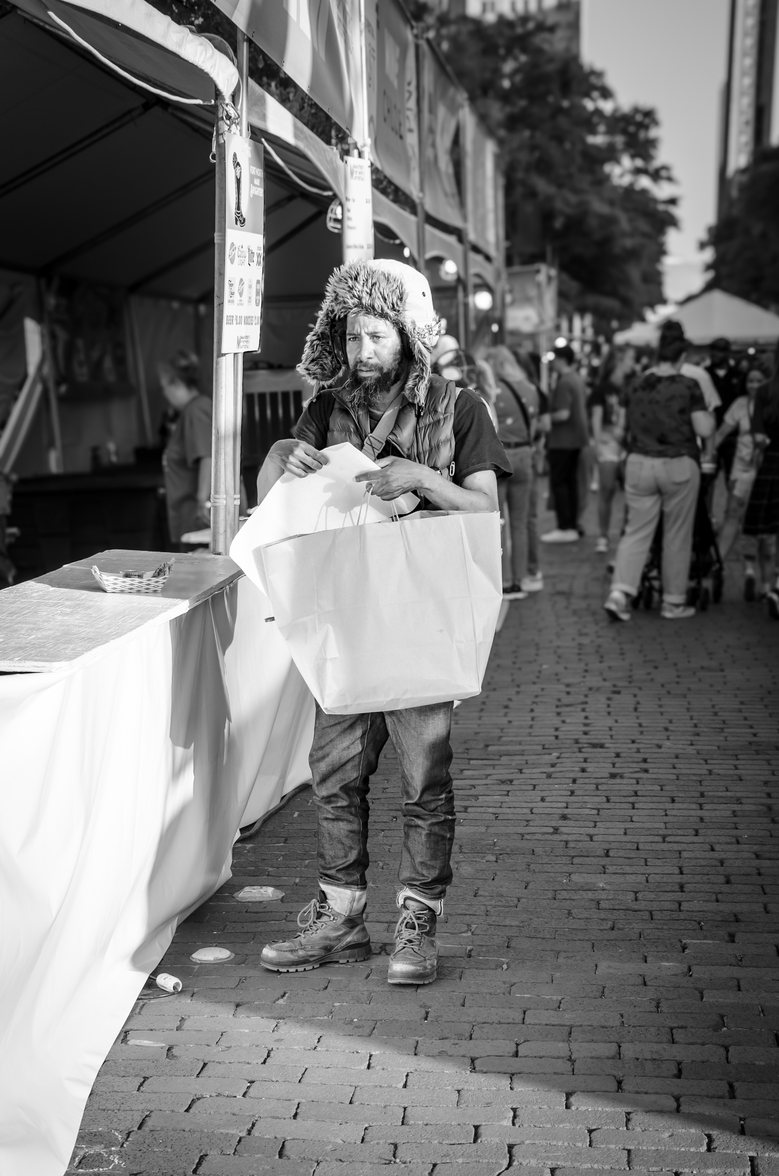 Caught in a sliver of light between movement and stillness, a man clutches a paper bag as if holding more than objects—perhaps expectations, survival, or silence. His heavy boots, fur-lined hat, and layered clothing suggest resilience.
