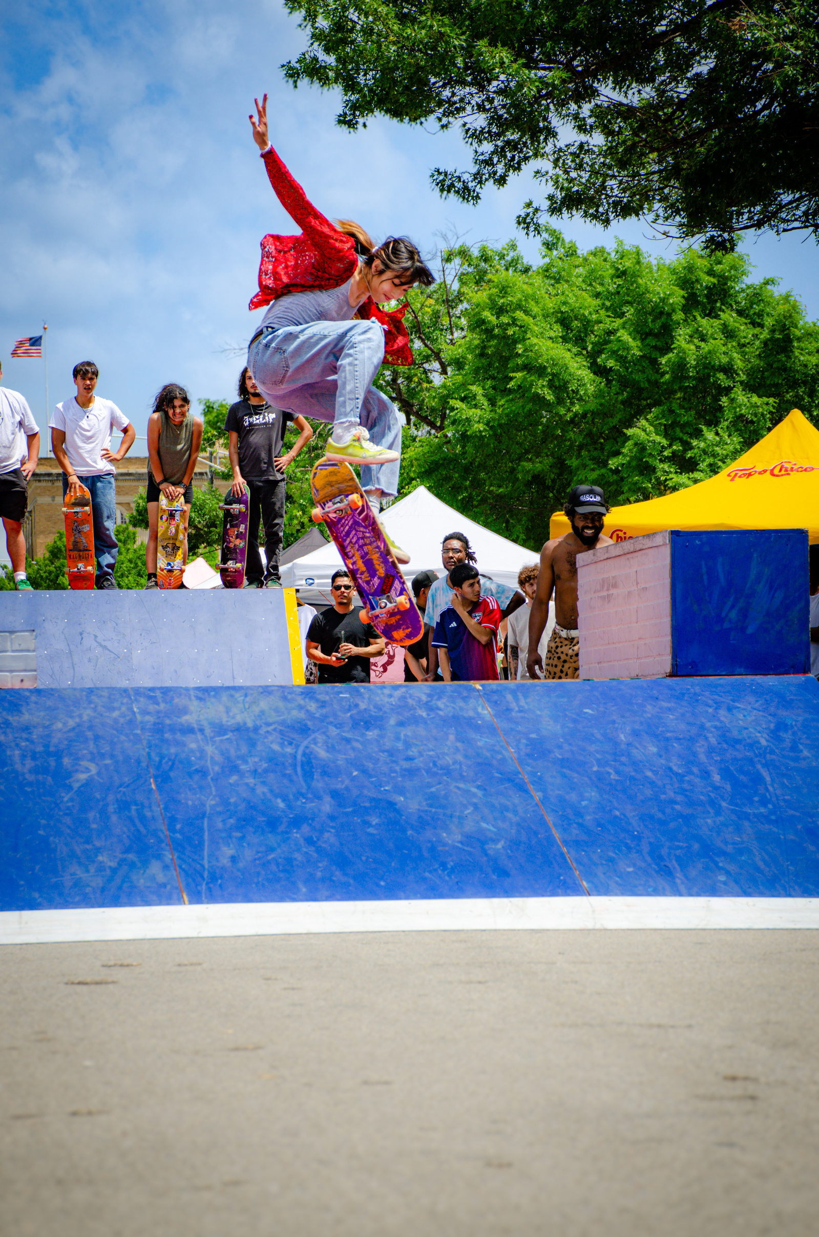 Mid-kickflip and mid-shout, the red-jacketed skater flashes like a match against the blue ramp. Eyes watch, hands lift, and she rises into the trick like a spell being cast.
