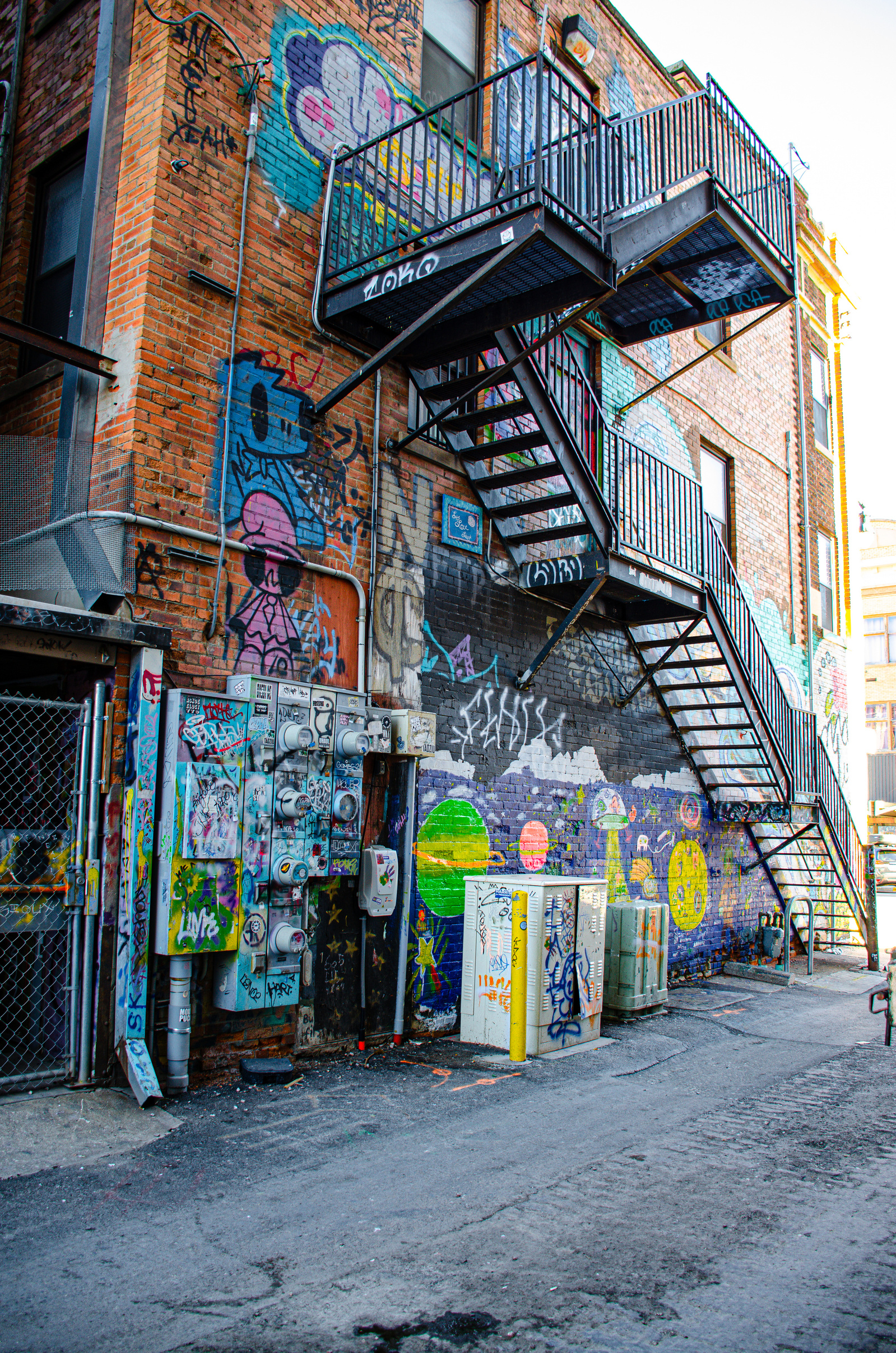 An electric nest of meters and wires anchors the wall, while above and around them—planets, astronauts, symbols of escape. This is the contradiction of the alley: tethered to function, dreaming in color.