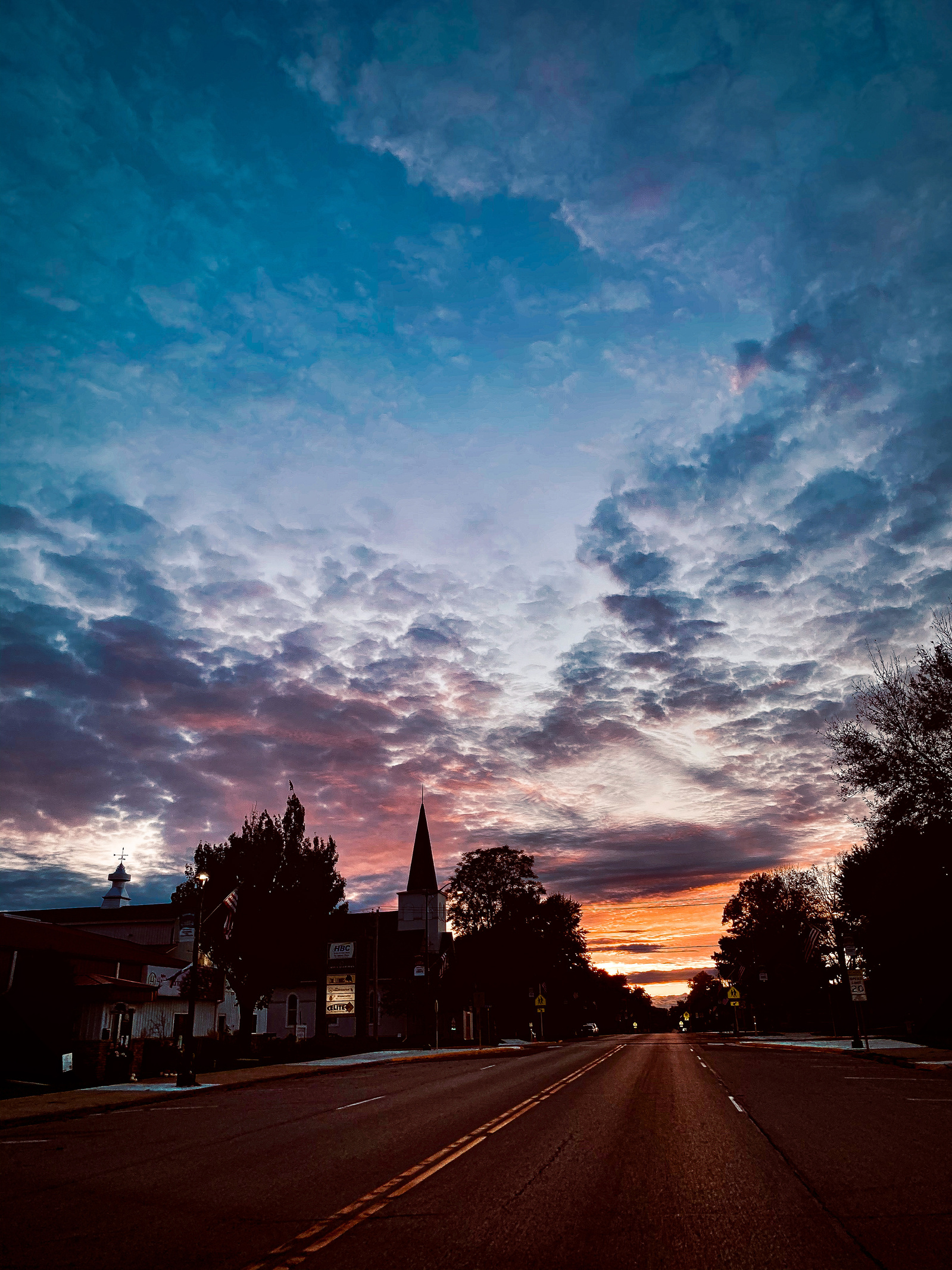 Church spires pierce a tapestry of clouds ablaze with sunset hues, as small-town America settles into evening. The empty street leads eyes toward a horizon painted in natures most vivid palette, celebrating the quiet beauty of rural dusk.