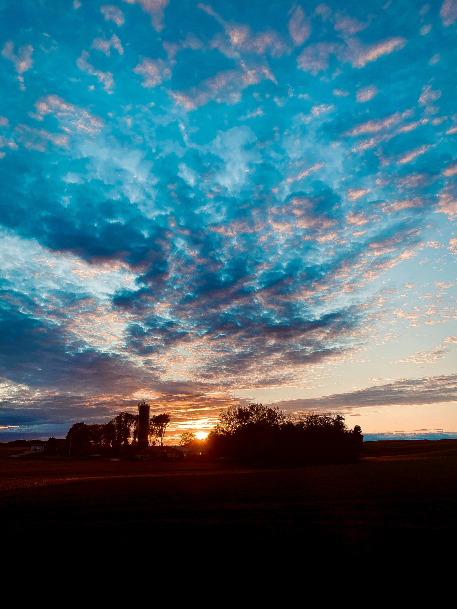 A lone silo stands watch against an explosive sky, where clouds scatter like embers across azure depths. The pastoral scene captures the dramatic interplay between human presence and natural grandeur, framed by the days final light.