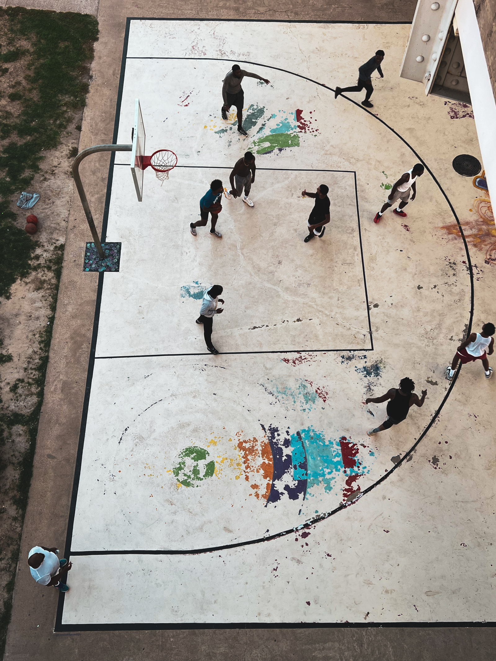 Aerial view of a community basketball game—movement, sweat, and rhythm play out on a court faded with time and color. The peeling paint echoes the legacy of every game ever played here, unrecorded but remembered in bodies and breath.