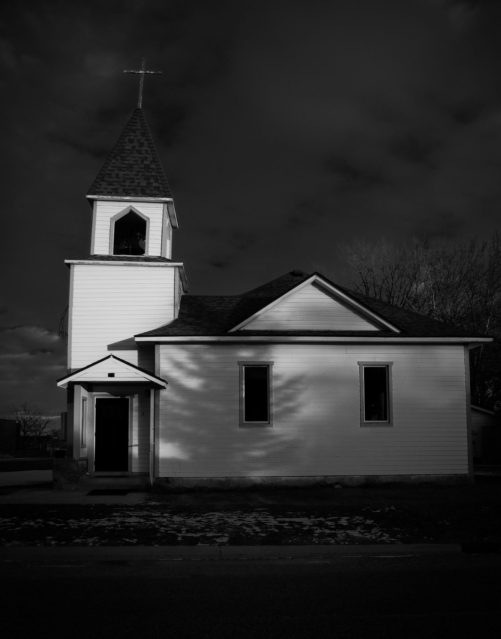 White clapboard church at dusk, windows dark, dramatic sky. The last light of day slips away, leaving the structure in contrast—its door ajar, its silhouette sharp. Faith doesn’t always gleam; sometimes, it broods.