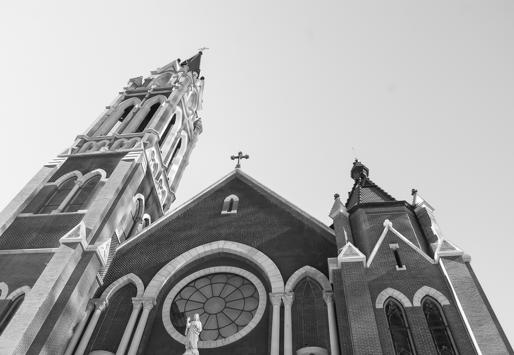 Gothic brick church with high steeple and statue below circular stained glass. The spire reaches into a pale sky, a vertical prayer in brick and glass. Below, a solitary statue gazes outward—not as sentinel, but as witness.