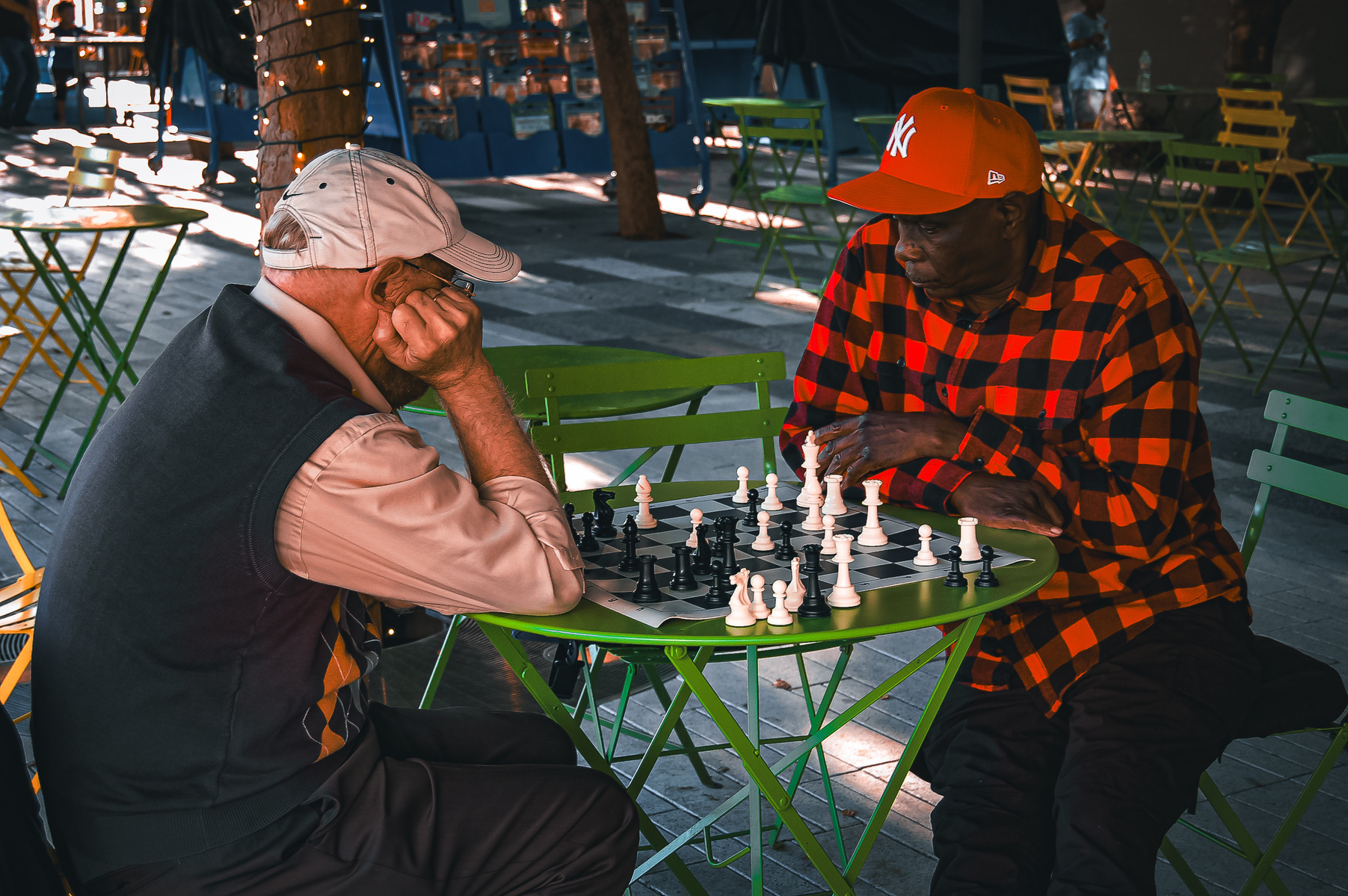 Two men play chess beneath soft café lights, locked in thought. Time slows between moves. Their differences fade in the ritual of strategy, concentration, and quiet companionship.
