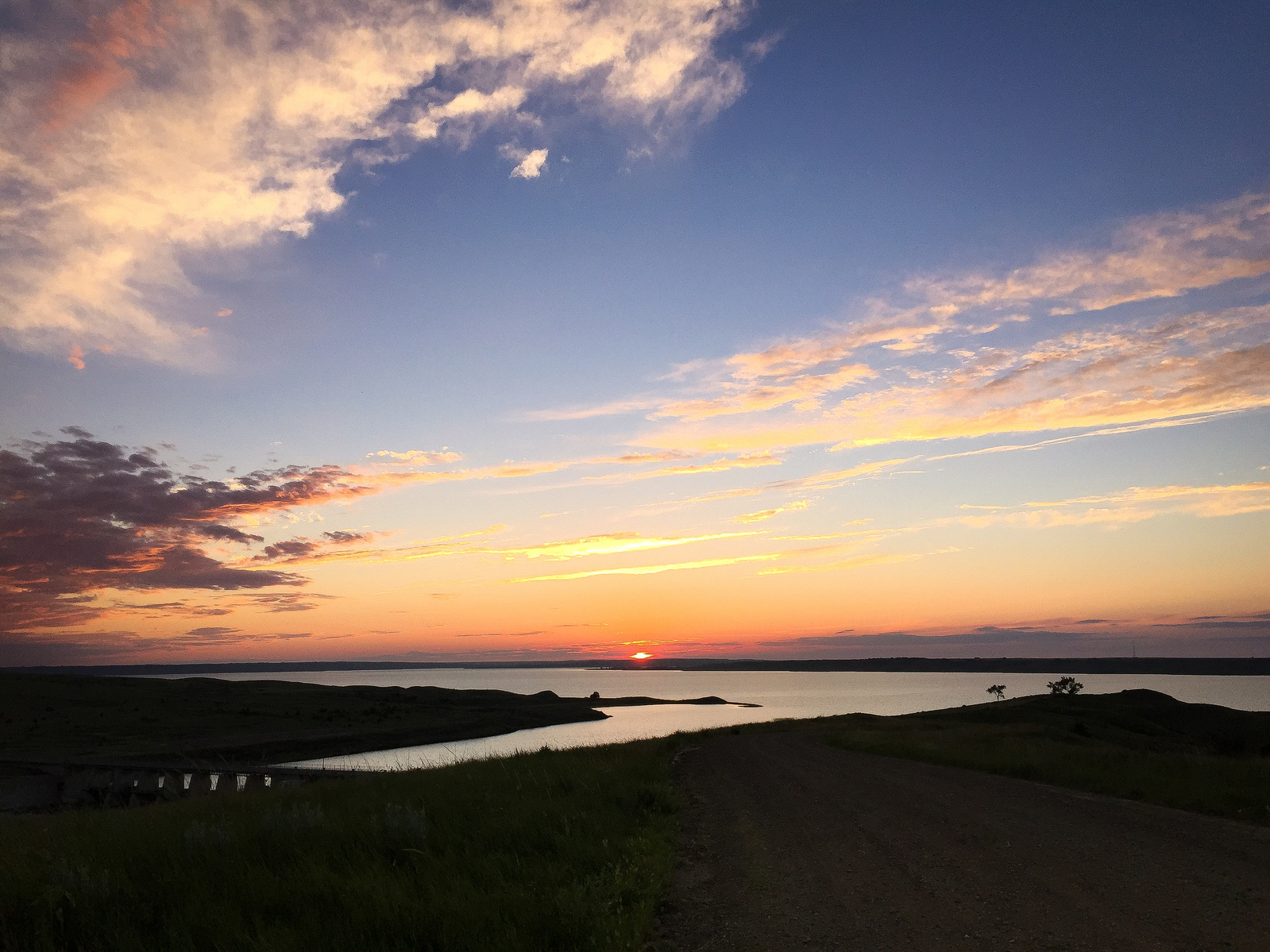 Evening light paints the sky in layers of amber and gold, reflecting across the still waters while silhouetted hills create a natural frame. The composition draws viewers through winding paths to the horizon, where sun meets water in a moment of perfect.