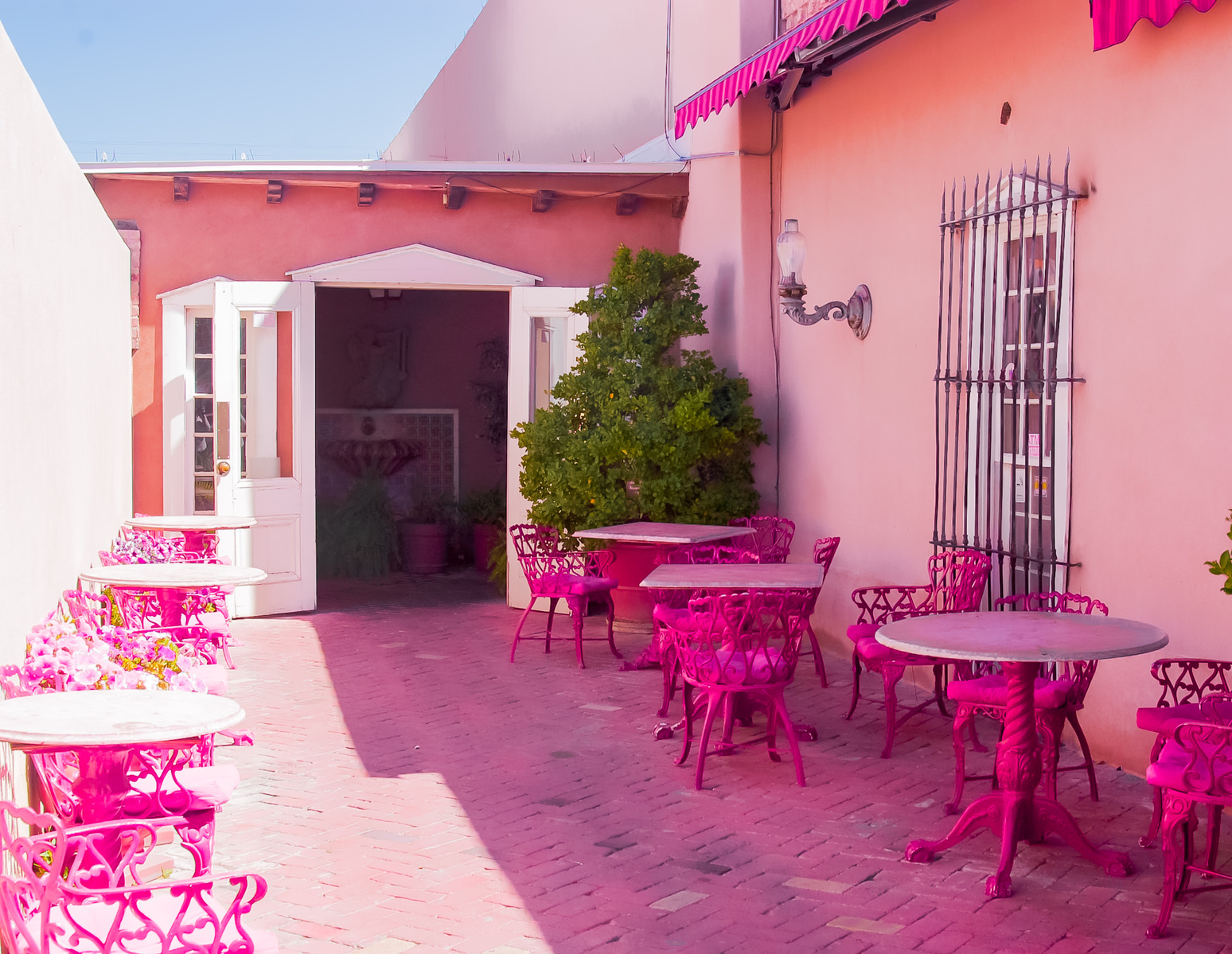 A courtyard drenched in unapologetic color—where everything feels paused in warmth and whimsy. The bold pink tables and chairs almost hum under the afternoon sun, inviting a slower moment, a quiet indulgence.