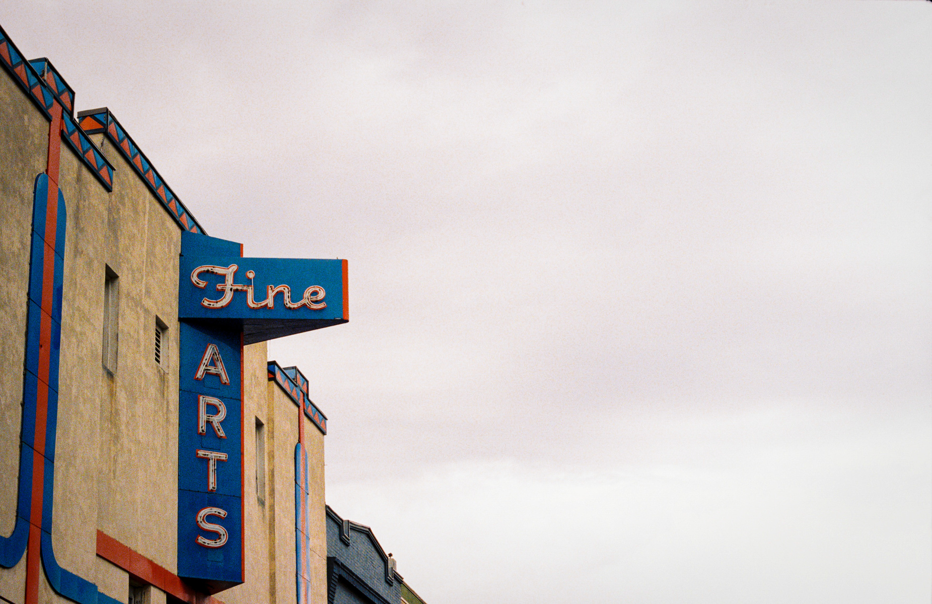 A lone sign juts into a washed-out sky, its script curled like a forgotten invitation. Once the promise of escape, of velvet curtains and silver screens, it now marks the border between memory and architecture.