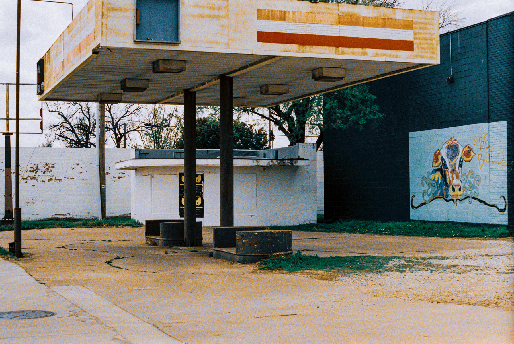 A sun-bleached gas canopy towers over cracked pavement and bold mural art—an intersection of abandonment and expression. The slogan says Be Bold, and the scene dares you to see beauty in the forgotten.