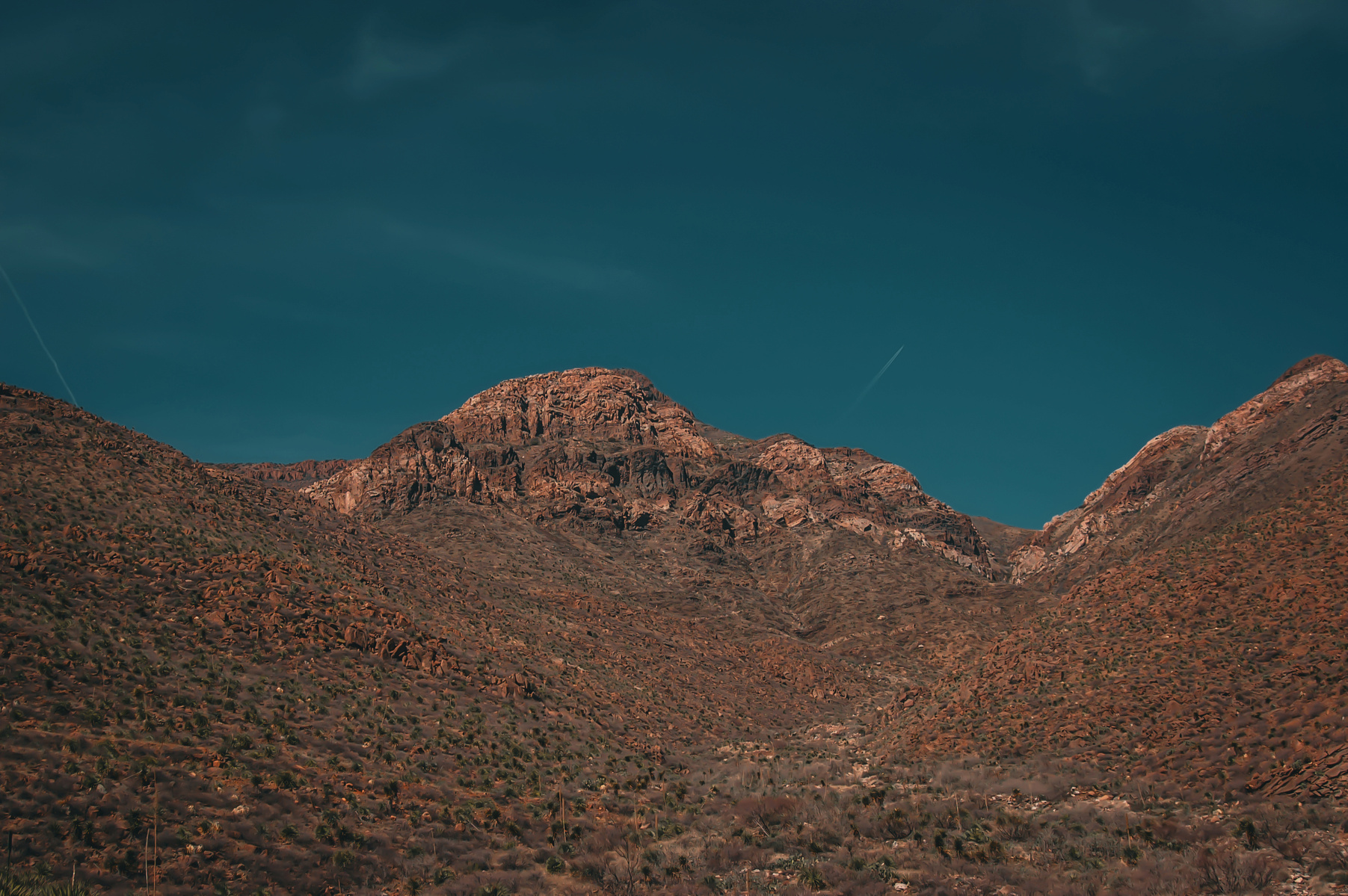 The mountain rises without asking to be followed, its slopes carved by time rather than purpose. Shadows drift between the ridge line like forgotten paths, while two contrails stretch thinly across the sky—vanishing lines that point nowhere in particular.