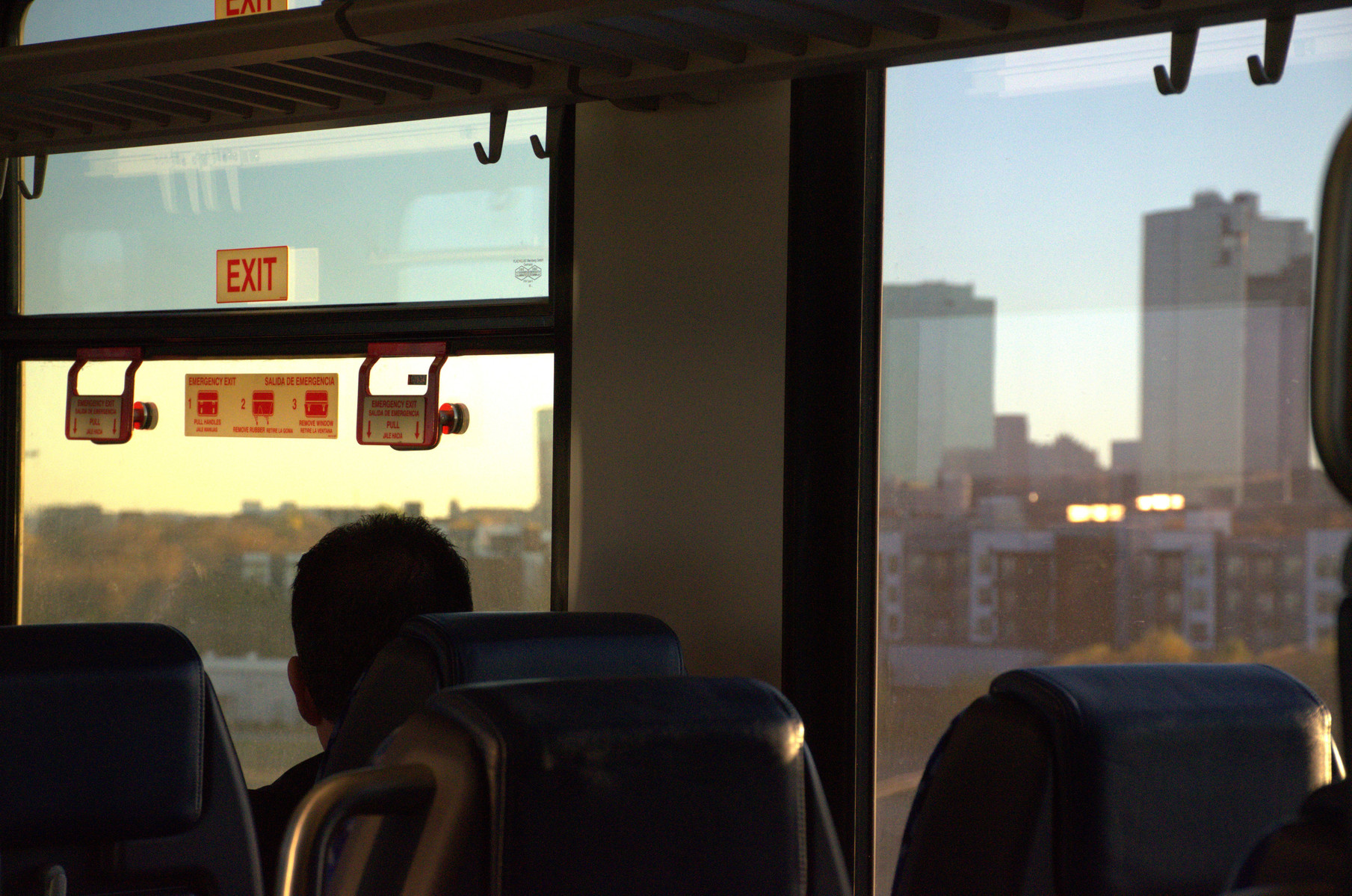 A lone commuter stares out the window as golden light slips across the city skyline. The train hums forward, but his thoughts stay behind. Caught between movement and memory, he becomes a silhouette in the fading day — part of the view, not the journey.