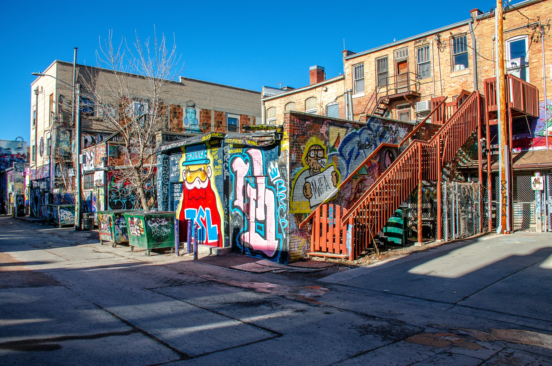 Sunlight spills over a wall of cartoonish defiance and layered commentary—faces, letters, stickers, memories, all shouting quietly into the alley’s hush. This is where the noise lingers long after the crowd has left.