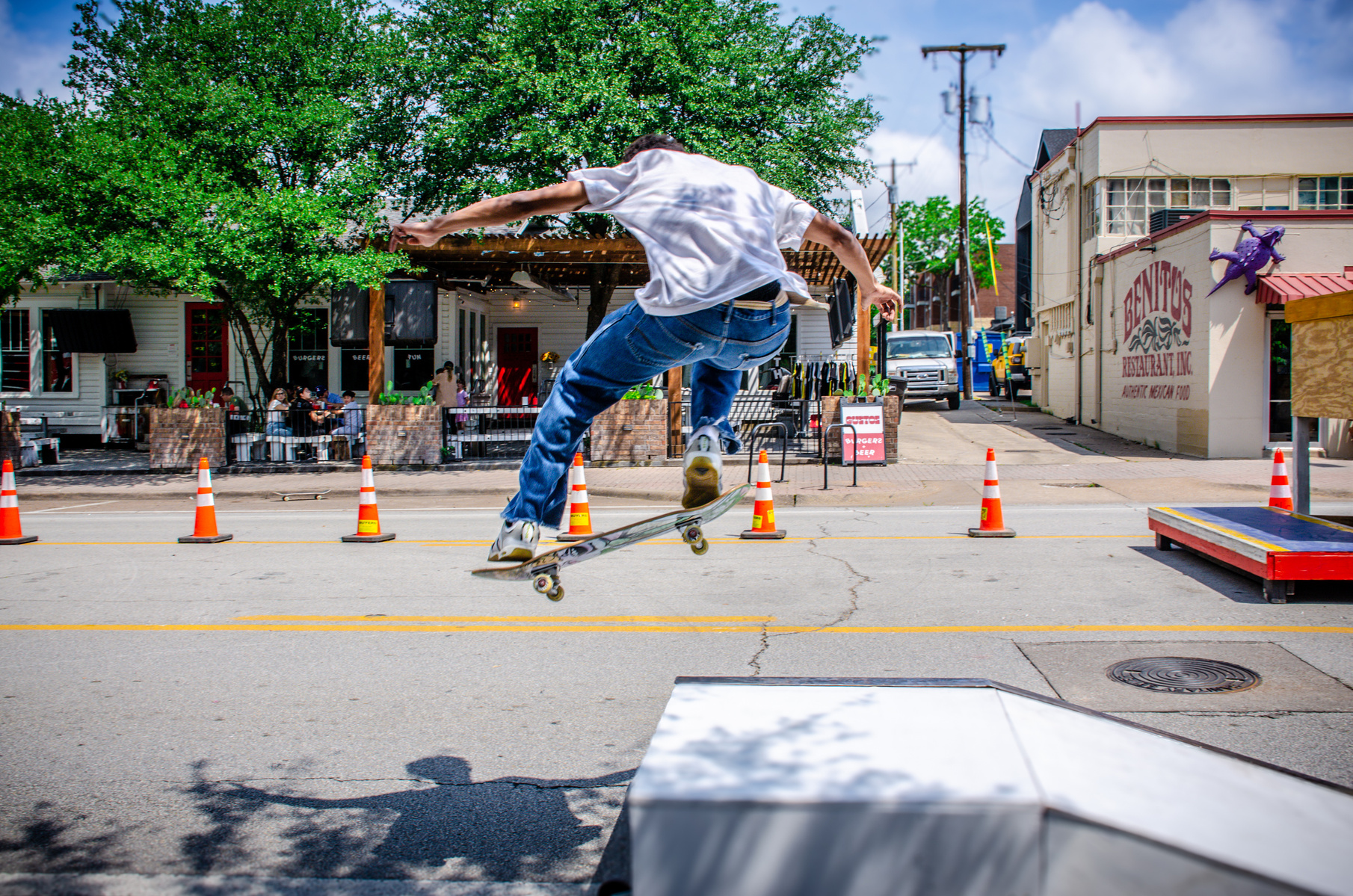 A fully committed flight—knees bent, shirt whipping, shadow stretched across the street. The crowd is still, the board is not. This is what it looks like to meet the air with intent.