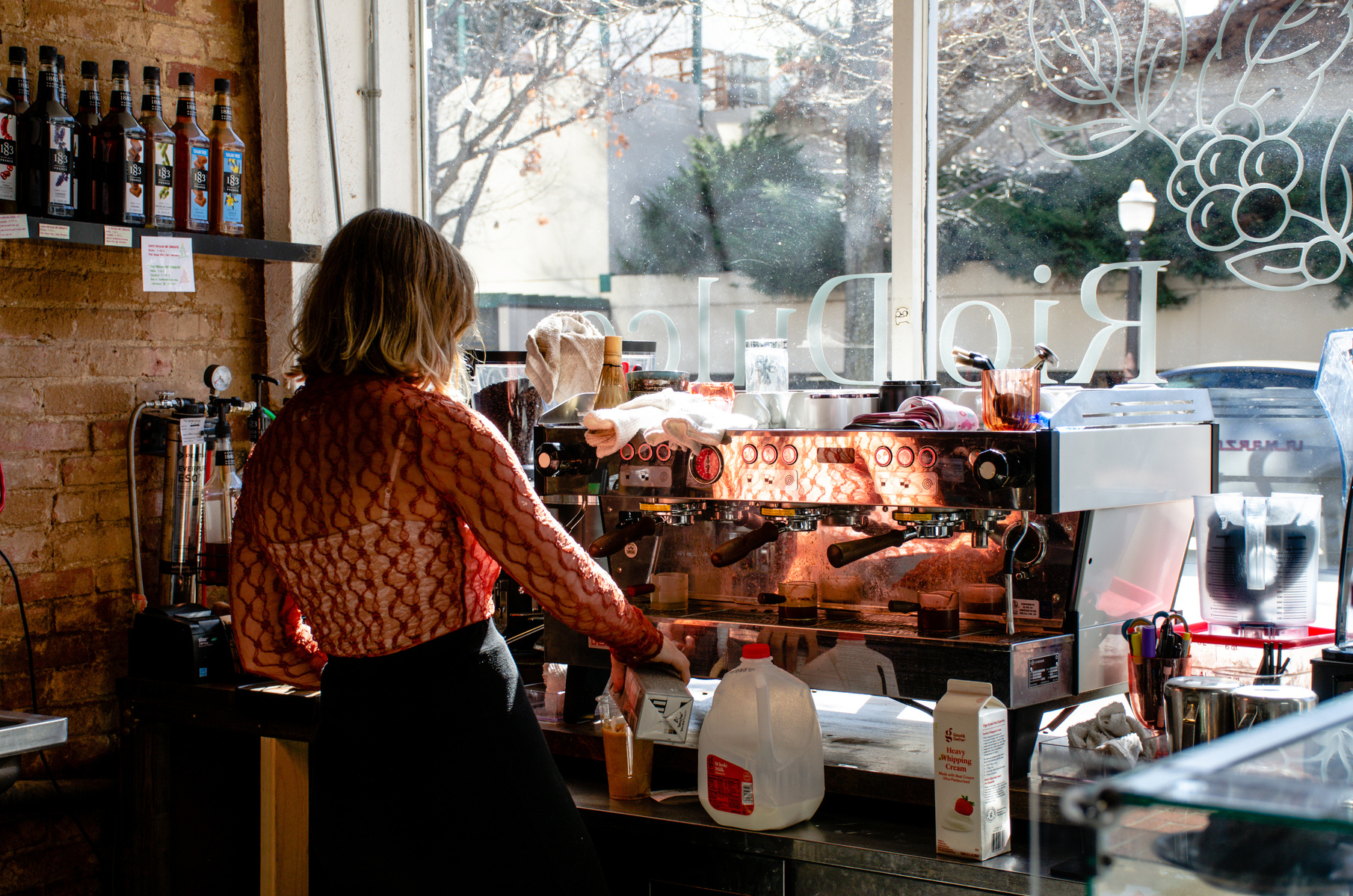 A barista works the espresso machine, morning light pouring through the café window like steam. Her posture and the cluttered counter reflect the quiet yet often overlooked art of repetition, of crafting care into every cup.