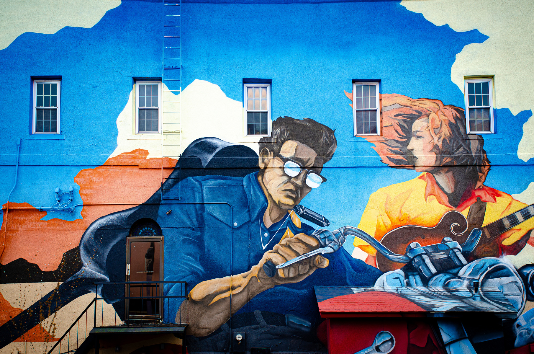 A collision of motion and music, this mural channels the grit of American rebellion and the fire of creative drive. The mirrored sunglasses and strumming hands suggest two paths—one racing forward, the other rooted in rhythm.