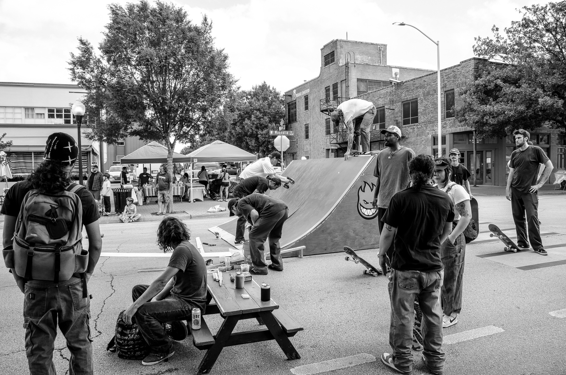 A moment stitched from bodies in motion and conversation. Elbows on knees, hands on tools, gazes tilted skyward or inward. The ramp is their altar; the asphalt, a fellowship of bruised knees and unspoken codes.