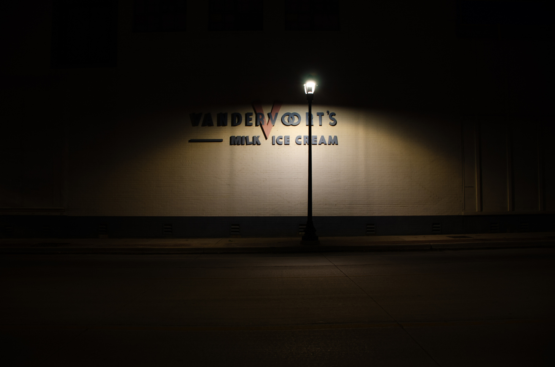 A streetlamp performs for no one, spotlighting a faded name. A brand remembered by architecture more than memory.