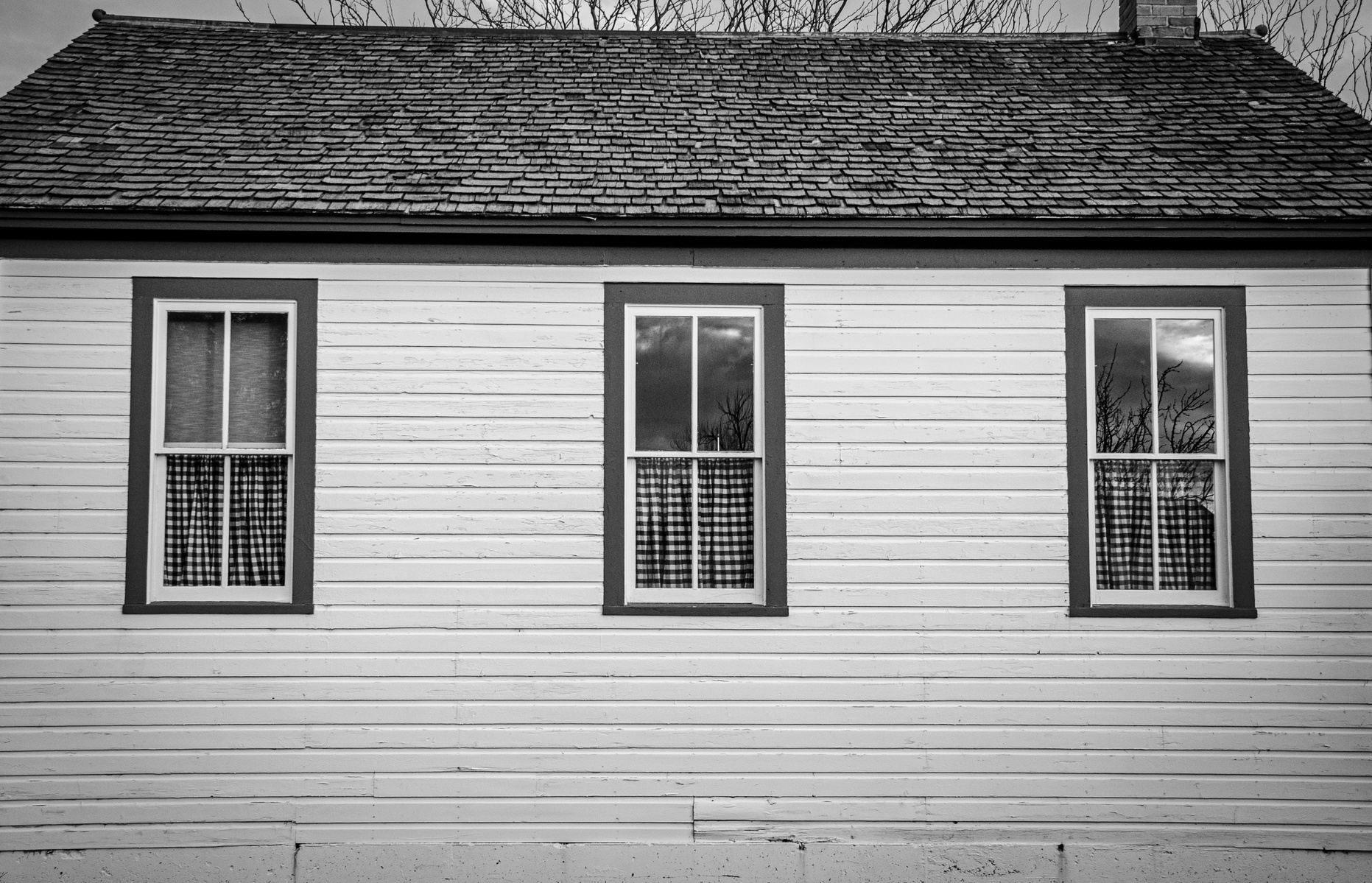 Symmetrical white wooden house or church with three windows and gingham curtains.
Every window is a mirror, every curtain a boundary. This façade, simple and unbothered, echoes a kind of spiritual domesticity—faith lived plainly.