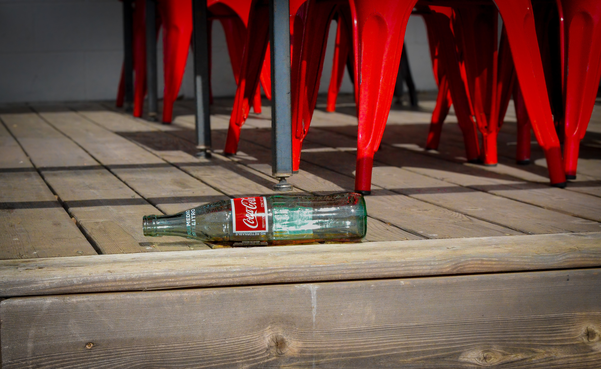 A half-liter Coke bottle rests on the edge of a day. Shadows stretch over the deck like a sigh. Someone was here. They left quietly.