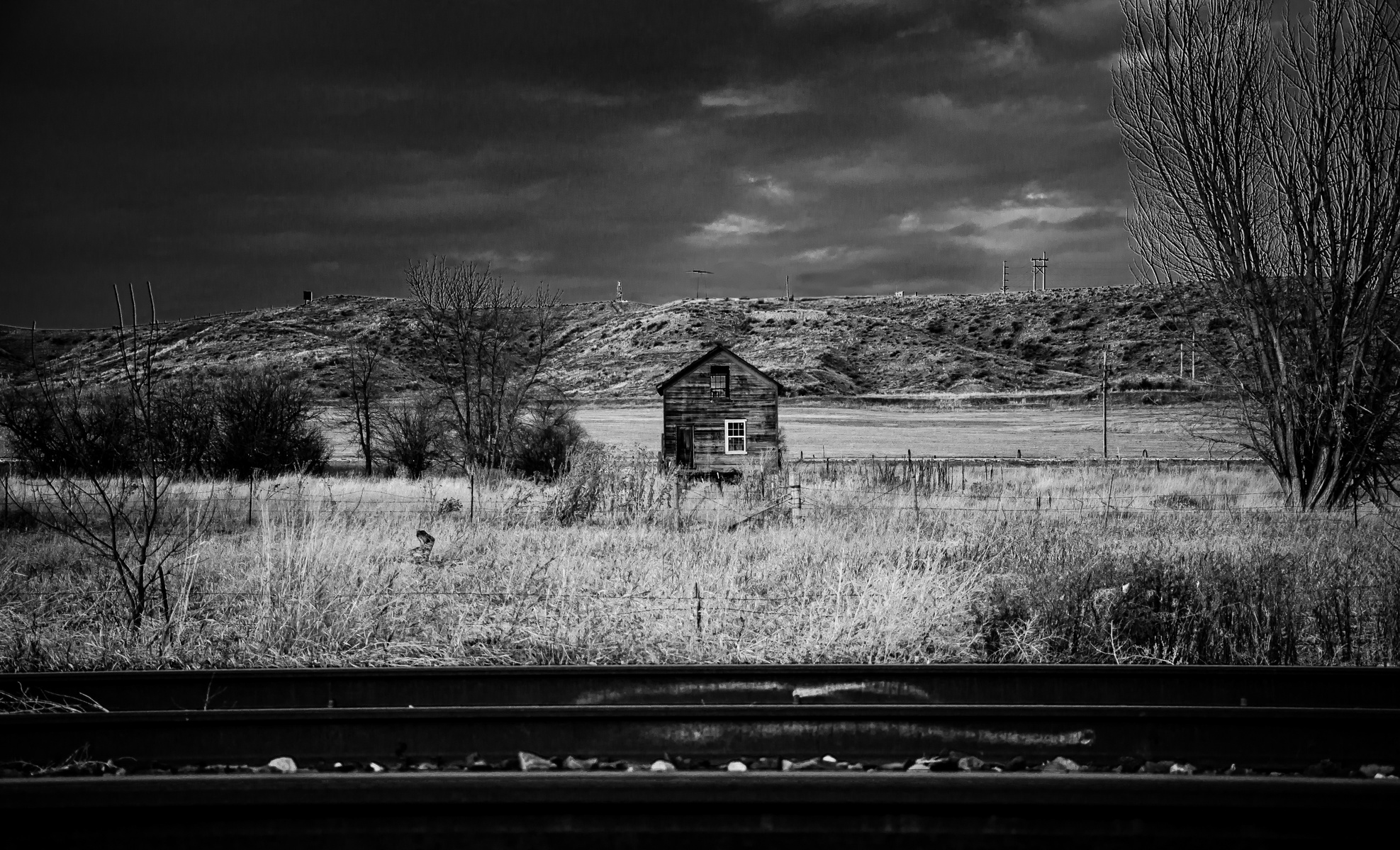 A lone wooden house stares back across brittle grass and silent rails. Abandoned or just waiting? This image holds a rural ache, a memory suspended in monochrome stillness.